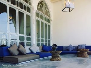 Picture of Hotel Efendi in Akko. The picture features one of the hotel's balconies. Low grey and blue sofa's and stone tables ornate the place and huge ottoman-style windows are facing the small chandelier and the marmer ground.