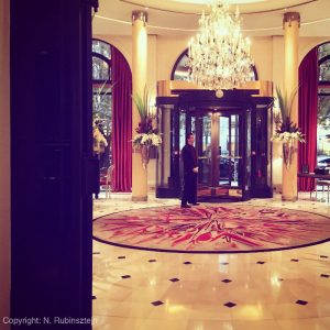 Picture of Hotel Plaza Athénée's entrance in Paris. The picture shows a bellboy, a huge chandelier, a round and Art Deco carpet, huge windows with red curtains and 2 Greek-styles pillars.