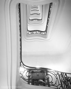 Black and white picture of Hotel Plaza Athénée in Paris featuring the beautiful staircase of the hotel. The first floor lead to a black door.