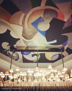 Picture of Hotel Prince de Galles in Paris featuring one of their magnificent chandeliers surrounded by a beautiful At Deco ceiling painting full of strong colors and forms.