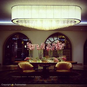 Picture of the Waldorf Astoria in Jerusalem. The picture features part of the hotel lobby with a huge white and chrystal chandelier, a huge wooden table with about ten pink flower bouquets. In front of the table are two light brown leather sofas.