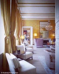 Picture of Hotel Dukes' Palace in Bruges showing the entrance to one of its lobbies. One can see purple and white sofas, beige curtains, hanging and standing chandeliers, a mirror, orange and yellow wallpaper. There is a door leading to the next lobby.