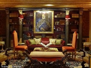 Picture of Gritti Palace in Venice featuring a cozy Renaissance living room. The walls are covered by chandeliers and the portrait of Venice's Doge, Andrea Gritti, painted by Titian. There are two orange chairs, a red sofa, beige pillows and a rug on the floor.